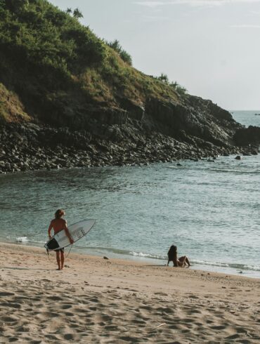 a surfer walking into the waters at West Nusa Tenggara, Indonesia with a surfboard on his side and a girl sitting on the seashore looking out in the horizon.