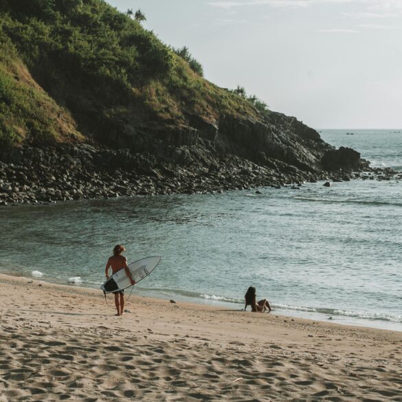 a surfer walking into the waters at West Nusa Tenggara, Indonesia with a surfboard on his side and a girl sitting on the seashore looking out in the horizon.