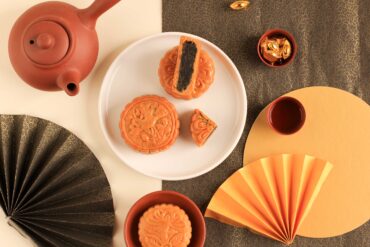 Traditional baked mooncakes served with Chinese tea in a Mid-Autumn Festival flat lay, featuring intricate gold fans, red teacups, and festive decor — perfect for celebrating the mooncake season in Singapore.
