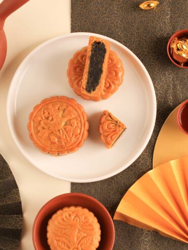 Traditional baked mooncakes served with Chinese tea in a Mid-Autumn Festival flat lay, featuring intricate gold fans, red teacups, and festive decor — perfect for celebrating the mooncake season in Singapore.