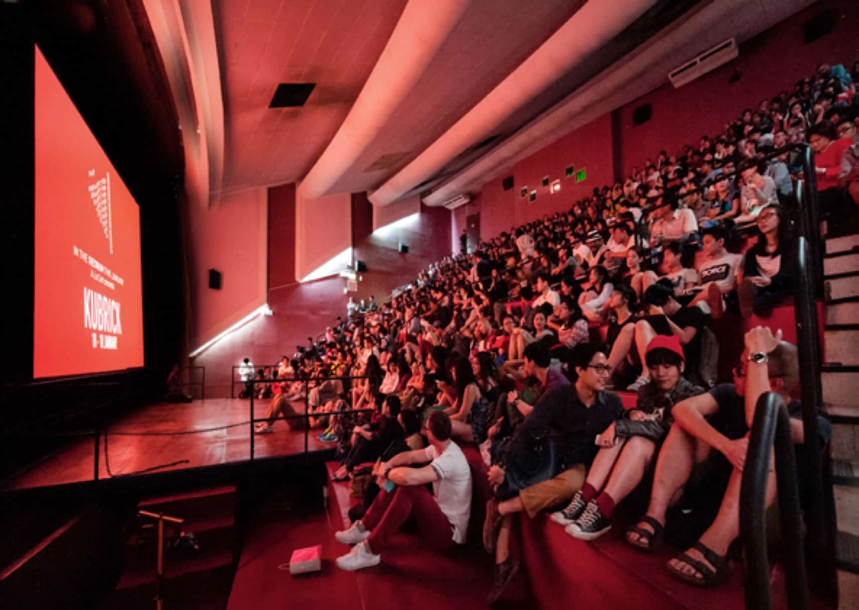A full house inside The Projector’s cinema hall, with audiences seated on tiered red steps watching a film screening on the big screen.