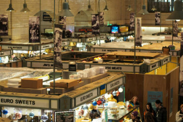 Indoor food market with vintage signage and diverse vendor stalls including Euro Sweets and fresh meats, bustling with shoppers under warm lighting.