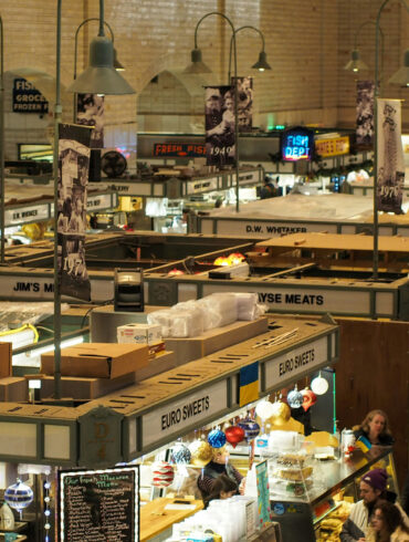 Indoor food market with vintage signage and diverse vendor stalls including Euro Sweets and fresh meats, bustling with shoppers under warm lighting.