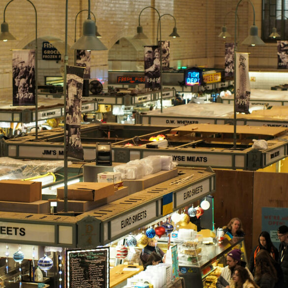 Indoor food market with vintage signage and diverse vendor stalls including Euro Sweets and fresh meats, bustling with shoppers under warm lighting.