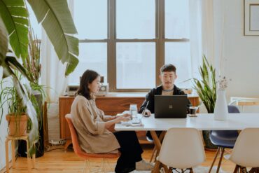 Two Asian digital nomads working remotely on laptops in a modern home office with natural light and indoor plants.