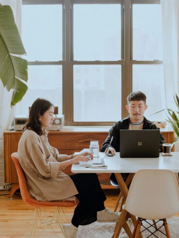 Two Asian digital nomads working remotely on laptops in a modern home office with natural light and indoor plants.