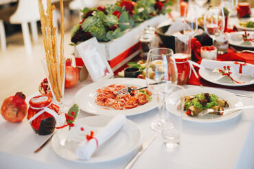 Elegant Italian Christmas Day lunch table setting with festive red-and-white decor, ribbon-wrapped napkins, fresh pomegranates, and pasta served with tomato sauce and grilled vegetables.