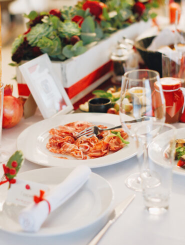 Elegant Italian Christmas Day lunch table setting with festive red-and-white decor, ribbon-wrapped napkins, fresh pomegranates, and pasta served with tomato sauce and grilled vegetables.