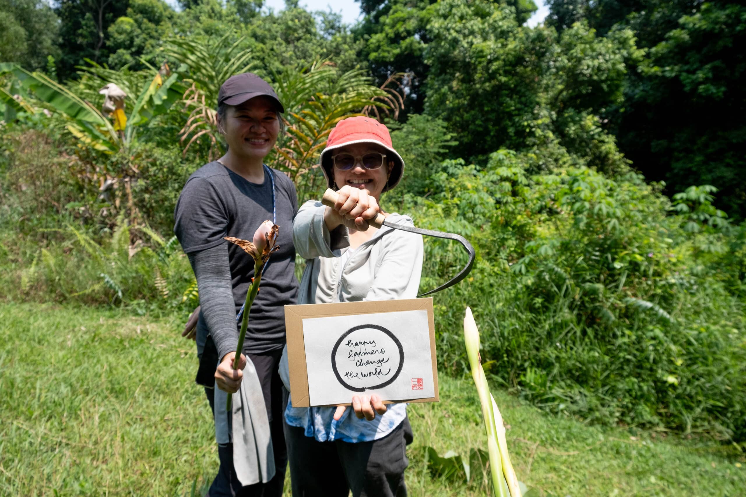 Two participants smiling in a lush, outdoor setting at the Living Earth Festival, holding harvested plants and a sign reading "Happy Farmers Change the World".