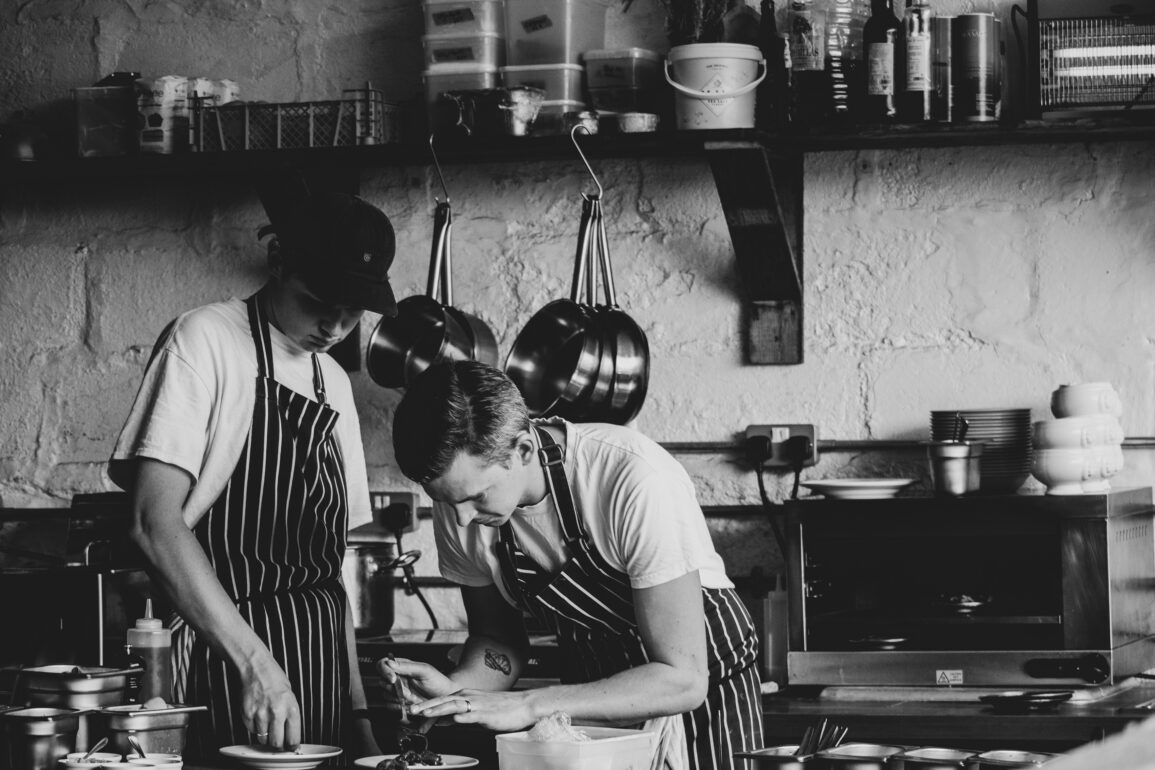 Black and white image of two chefs plating dishes in a rustic kitchen — showcasing hands-on culinary craft and teamwork in a modern restaurant.