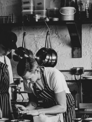 Black and white image of two chefs plating dishes in a rustic kitchen — showcasing hands-on culinary craft and teamwork in a modern restaurant.