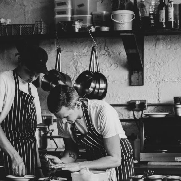 Black and white image of two chefs plating dishes in a rustic kitchen — showcasing hands-on culinary craft and teamwork in a modern restaurant.