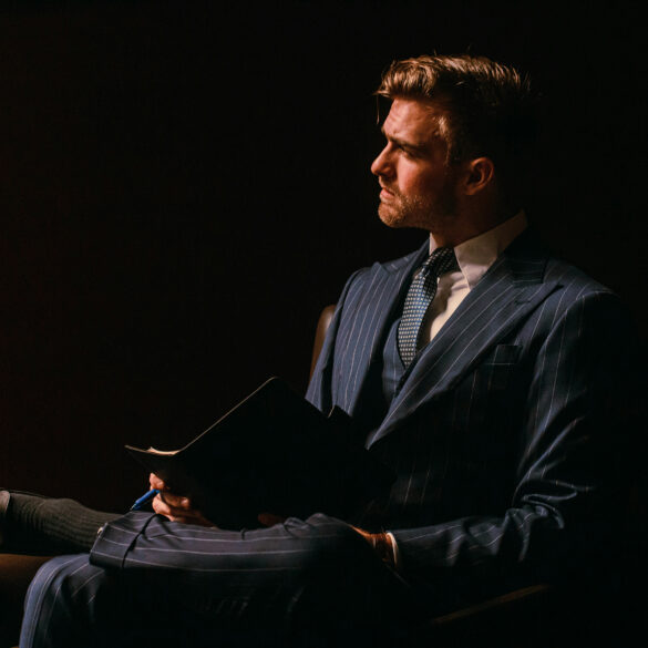 Man in a navy pinstripe three-piece suit sitting confidently in a dimly lit room, showcasing modern luxury businesswear for men.