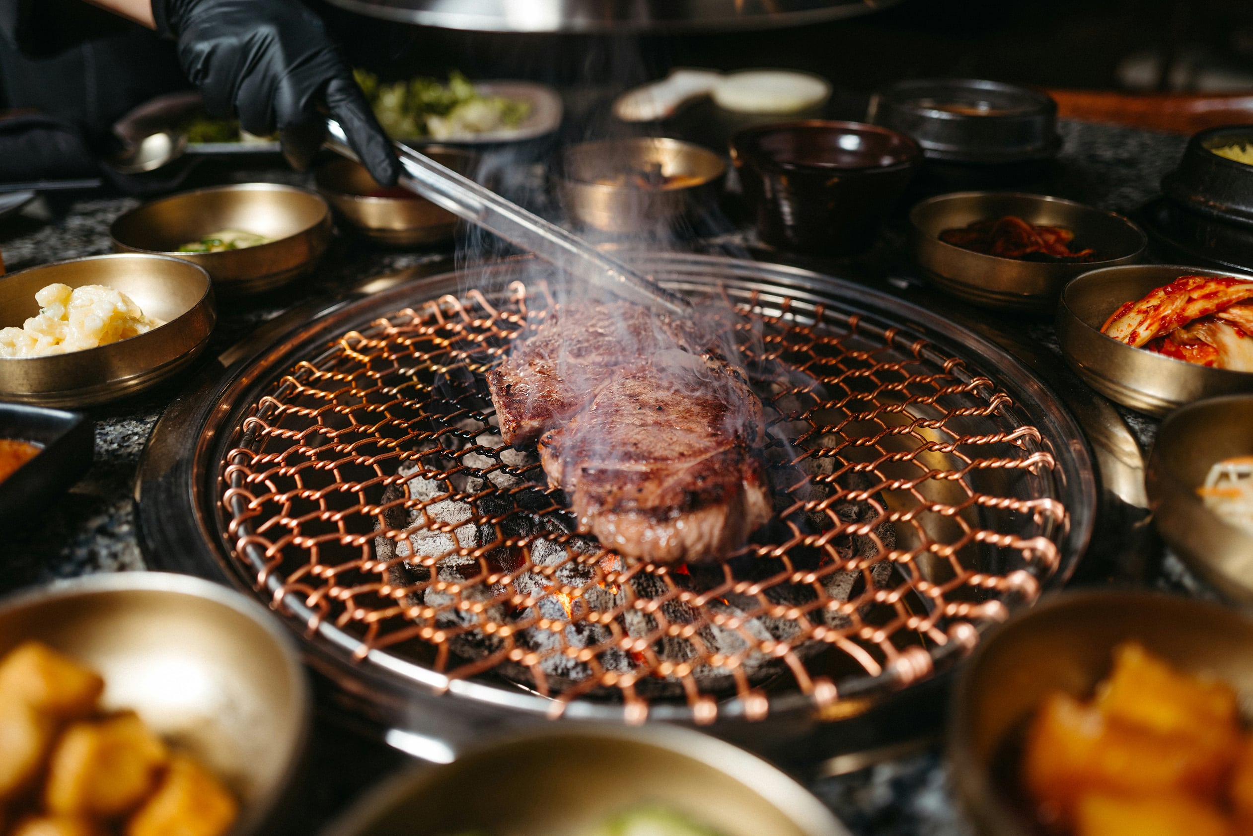 Korean BBQ steak grilling on a copper mesh over charcoal, surrounded by traditional banchan side dishes in golden bowls.