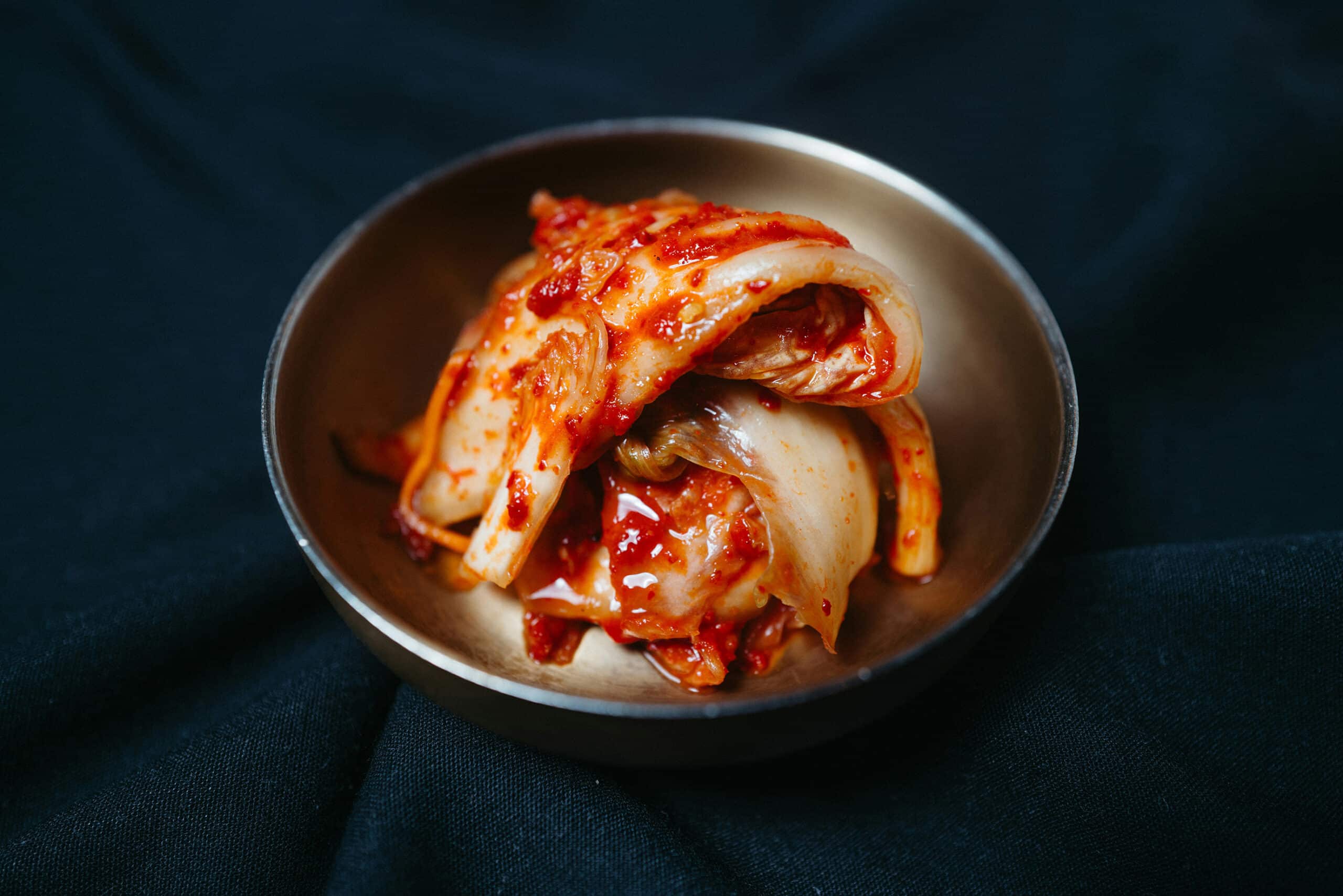 Traditional Korean kimchi made with napa cabbage and chili pepper, served in a brass bowl on dark fabric.