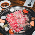 Thin slices of marbled beef grilling with vegetables at a Korean BBQ table, accompanied by white bowls of dipping sauces and banchan.