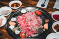 Thin slices of marbled beef grilling with vegetables at a Korean BBQ table, accompanied by white bowls of dipping sauces and banchan.