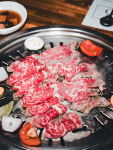 Thin slices of marbled beef grilling with vegetables at a Korean BBQ table, accompanied by white bowls of dipping sauces and banchan.