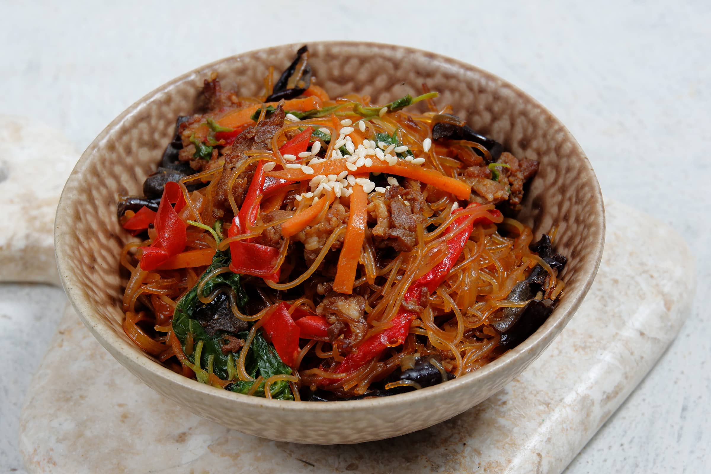 Korean japchae noodles with beef, vegetables, and sesame seeds in a rustic ceramic bowl on a light stone surface.
