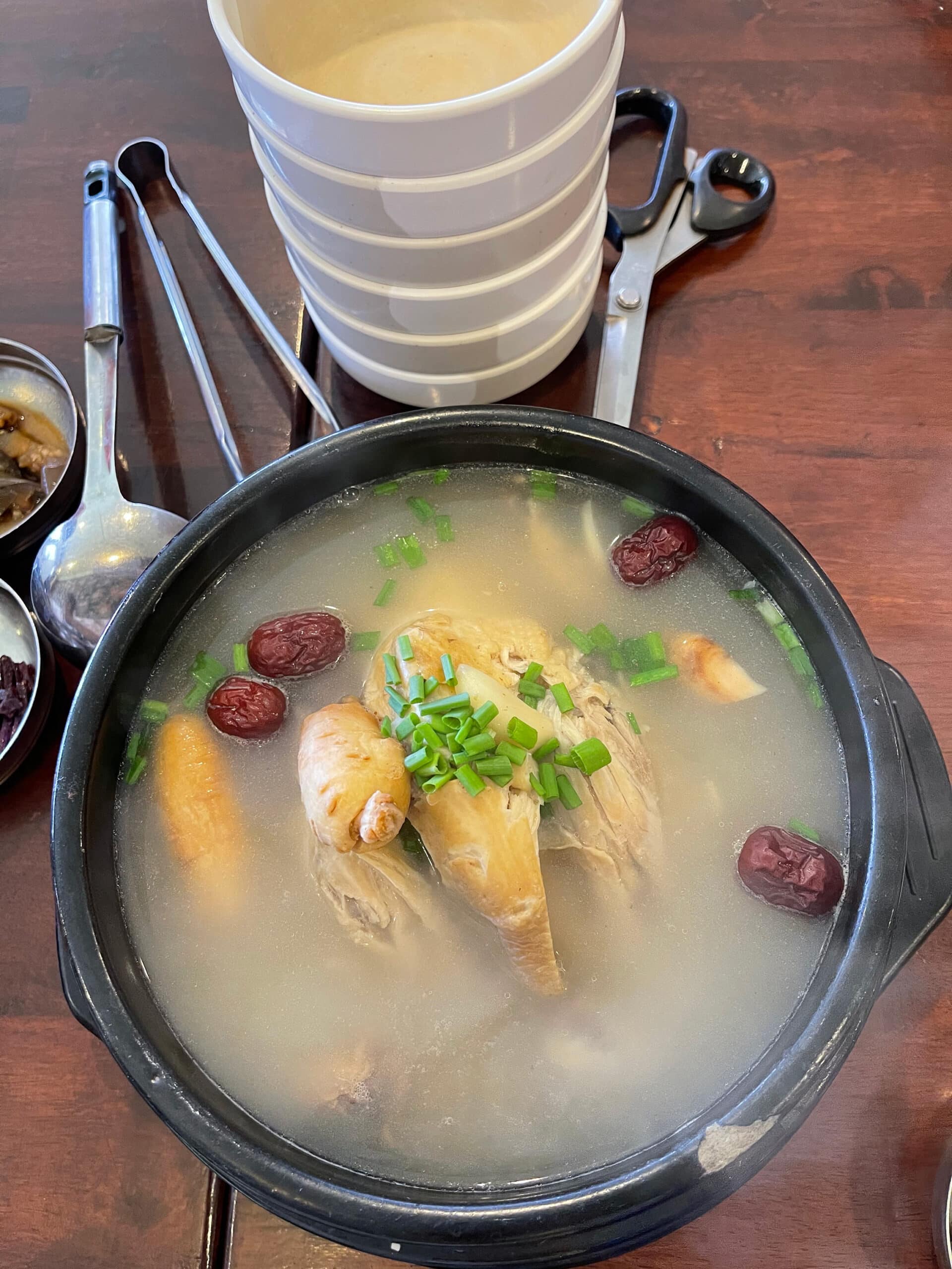 Traditional Korean samgyetang with a whole chicken, ginseng, jujube, and herbs in a hot stone pot on a wooden table.