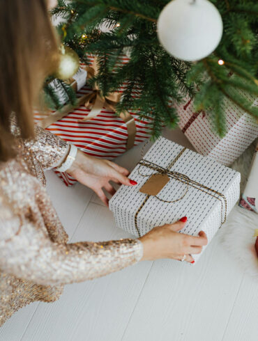 Festive Christmas scene with a woman in a sparkly dress arranging wrapped gifts under a decorated tree — representing joyful holiday gifting moments in 2025.