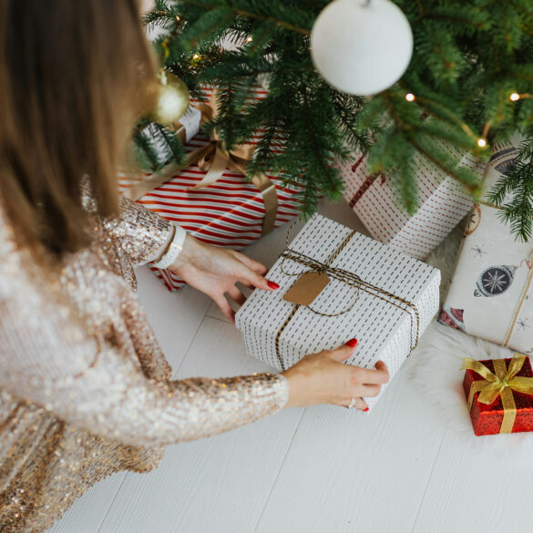 Festive Christmas scene with a woman in a sparkly dress arranging wrapped gifts under a decorated tree — representing joyful holiday gifting moments in 2025.
