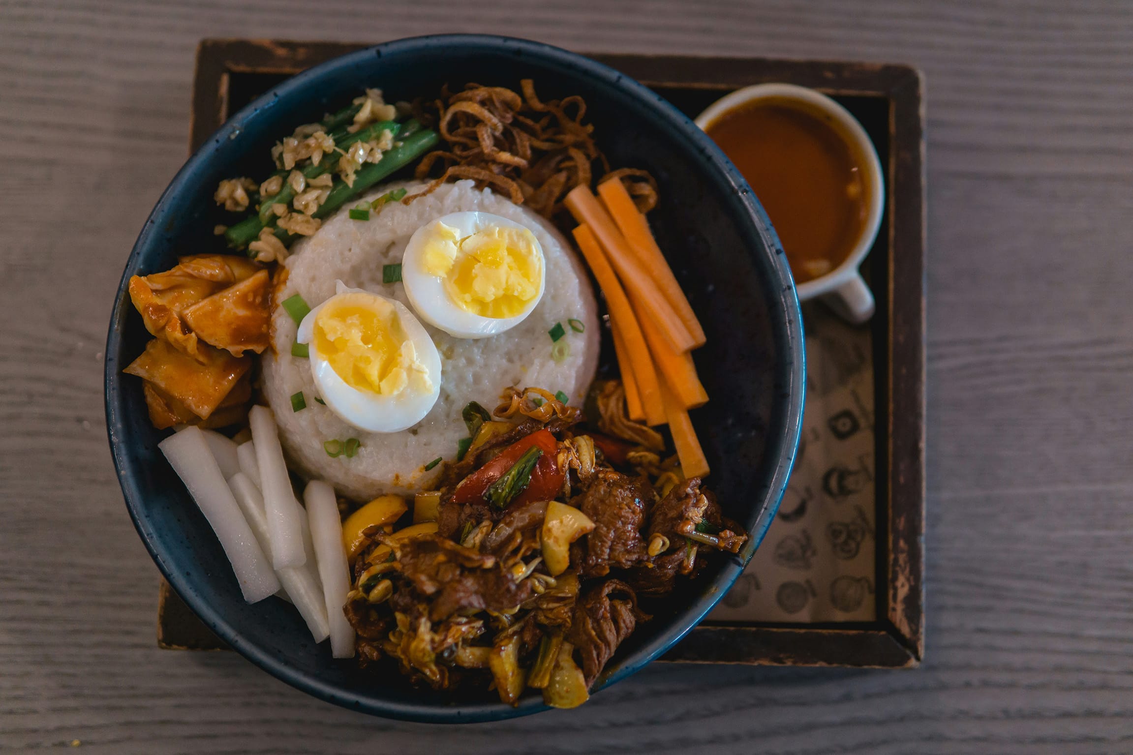 Korean bibimbap rice bowl topped with soft-boiled eggs, sautéed vegetables, pickles, and meat, served with gochujang sauce.
