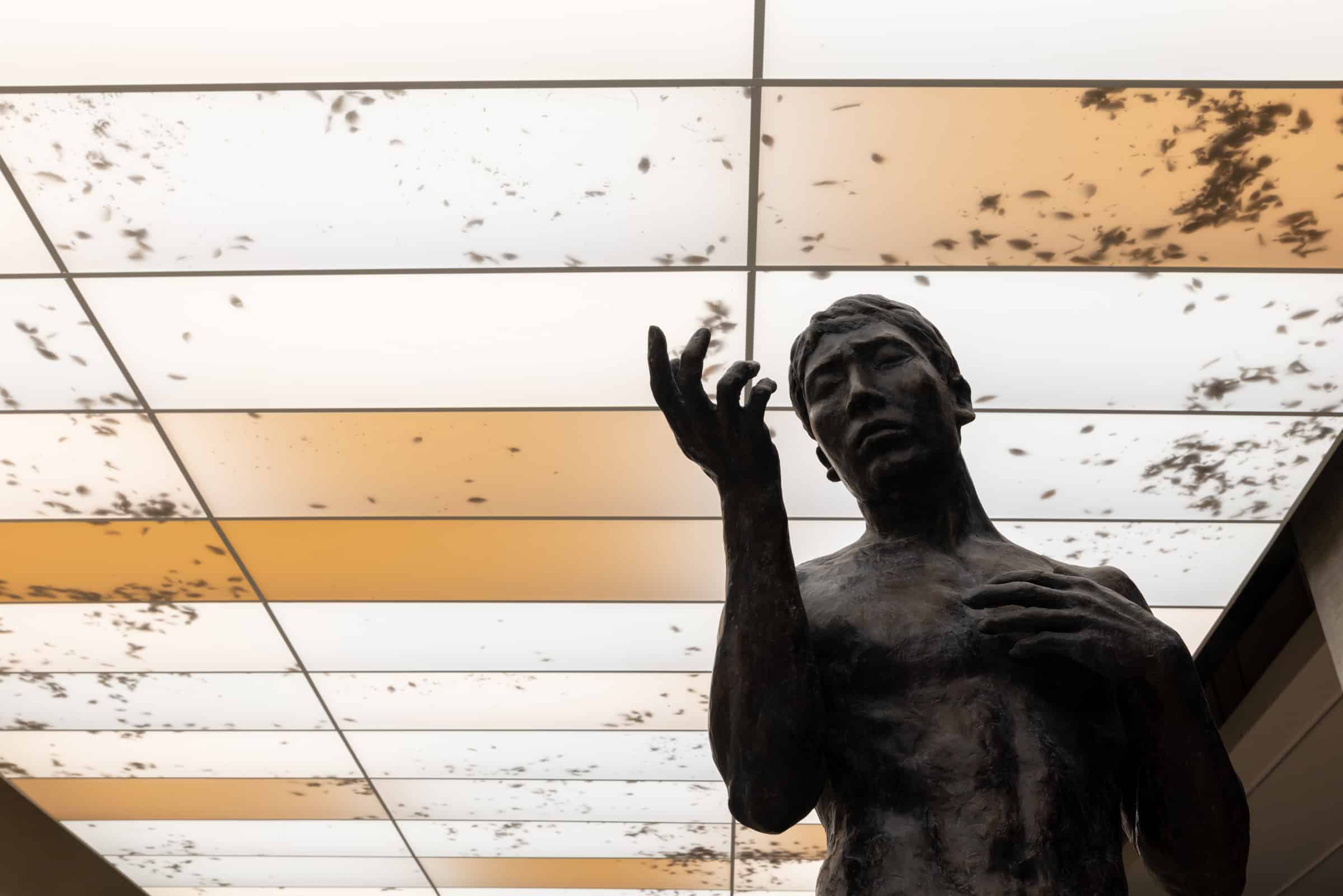 Bronze sculpture of a male figure gazing upward with one hand raised, set against a glowing ceiling with scattered shadow patterns, part of Álvaro Urbano’s TABLEAU VIVANT (A Stolen Sun) at Taipei Biennial 2025.