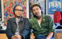 An Asian artisan couple wearing denim and military outfits sit side by side in a design studio, surrounded by colorful artwork and handcrafted goods