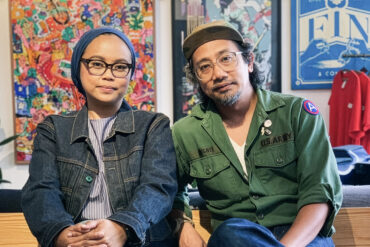 An Asian artisan couple wearing denim and military outfits sit side by side in a design studio, surrounded by colorful artwork and handcrafted goods