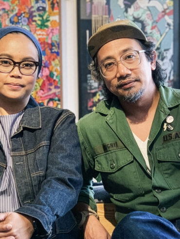 An Asian artisan couple wearing denim and military outfits sit side by side in a design studio, surrounded by colorful artwork and handcrafted goods