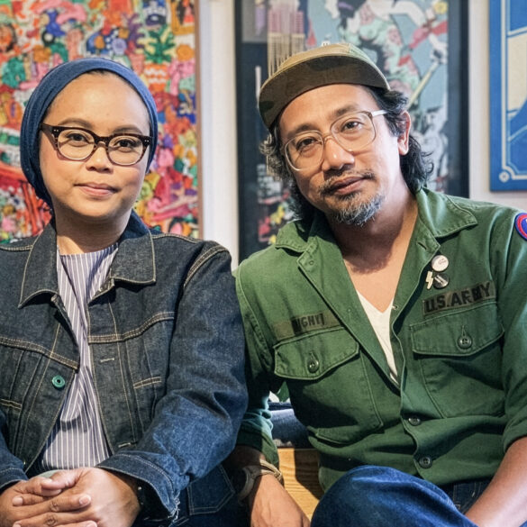 An Asian artisan couple wearing denim and military outfits sit side by side in a design studio, surrounded by colorful artwork and handcrafted goods