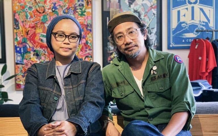 An Asian artisan couple wearing denim and military outfits sit side by side in a design studio, surrounded by colorful artwork and handcrafted goods