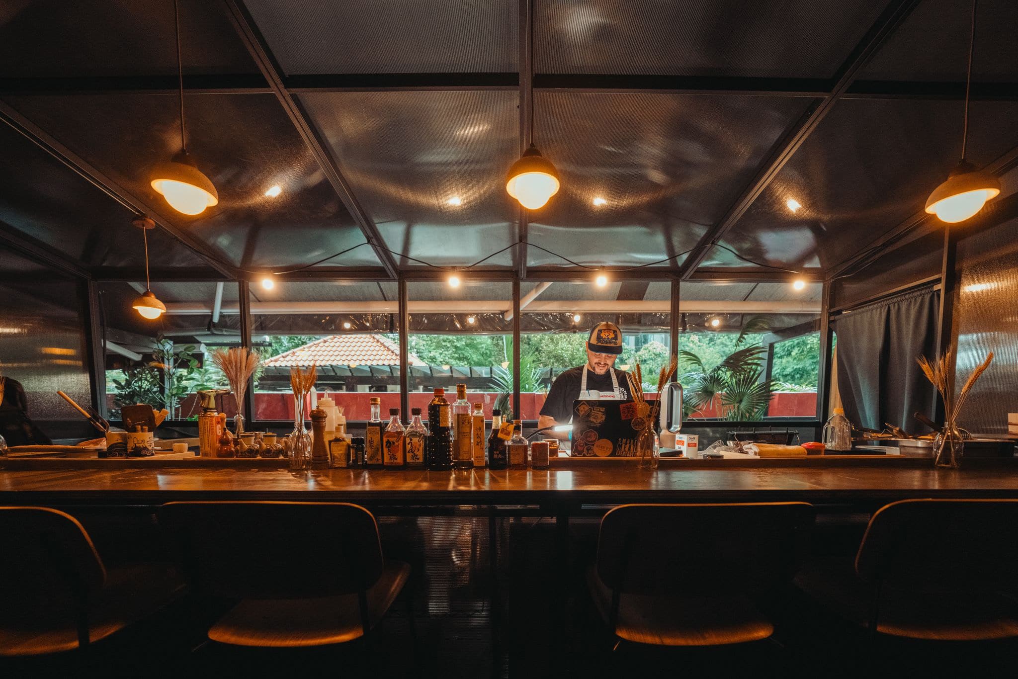 Interior of Jellyfish Sushi's ten-seat counter at Artichoke New Bahru with warm pendant lighting, chef preparing dishes, and view of tropical courtyard garden