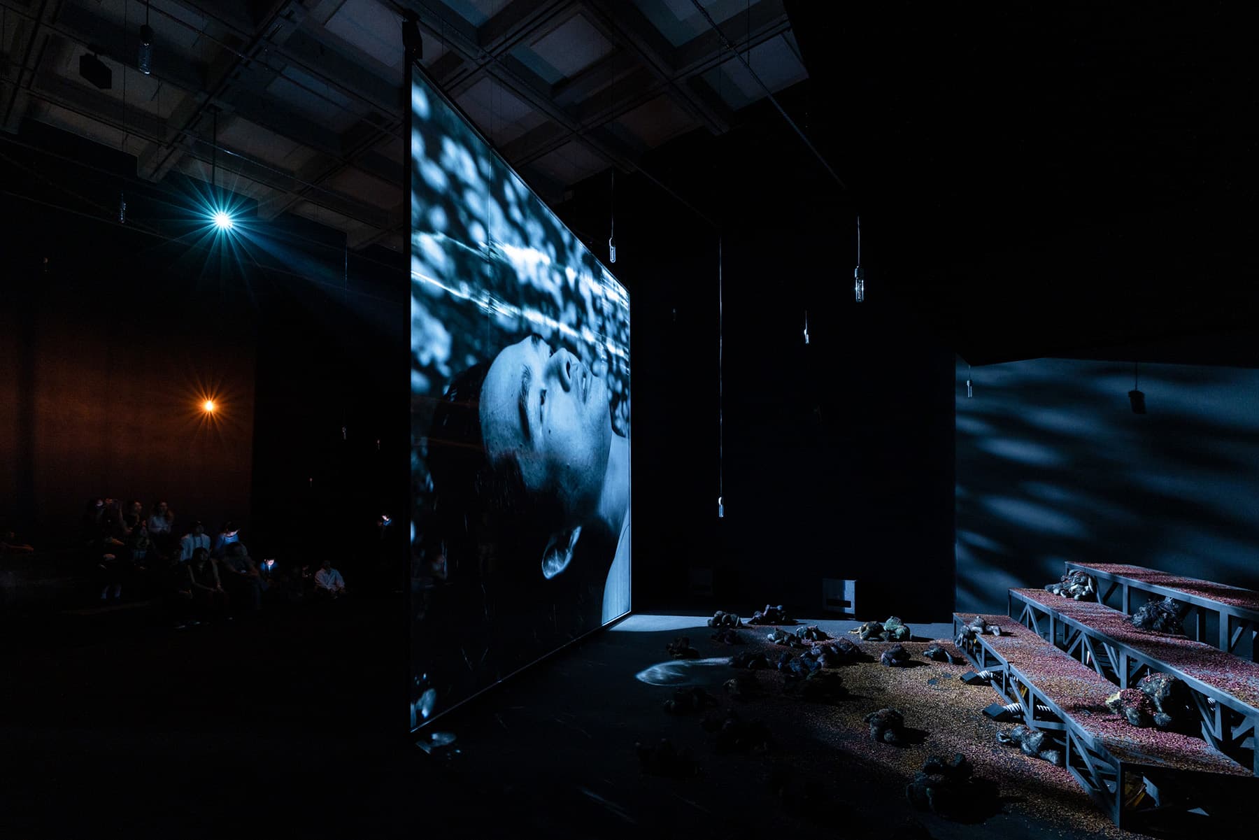Darkened installation space featuring a large video projection of a man’s face with moody lighting and scattered floral seating, part of Korakrit Arunanondchai’s Love after Death at Taipei Biennial 2025.