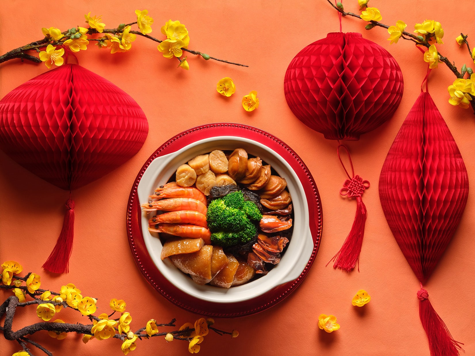 A top-down view of a traditional Chinese Pen Cai (treasure pot) filled with abalone, prawns, scallops, and broccoli, surrounded by red paper lanterns and yellow plum blossoms on an orange background.