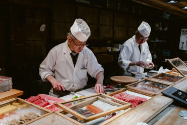 Japanese sushi chefs in traditional white uniforms preparing fresh fish at a sushi counter with wooden trays of tuna, salmon, and seafood for new restaurants in Singapore