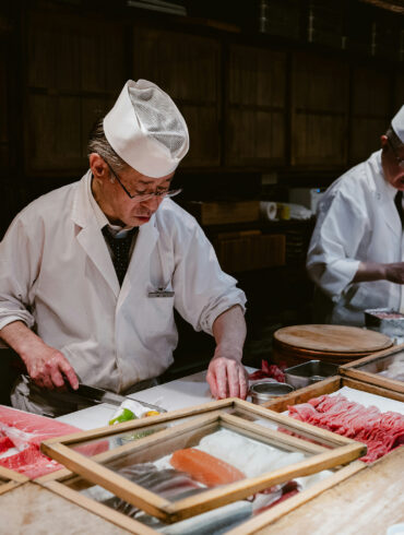 Japanese sushi chefs in traditional white uniforms preparing fresh fish at a sushi counter with wooden trays of tuna, salmon, and seafood for new restaurants in Singapore