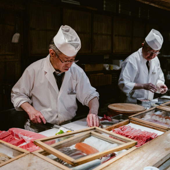 Japanese sushi chefs in traditional white uniforms preparing fresh fish at a sushi counter with wooden trays of tuna, salmon, and seafood for new restaurants in Singapore