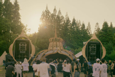 Golden hour at rural 2026 main stage with warm sunset glow and deep listening crowd immersed in techno, set against the forests of Fukushima, Japan