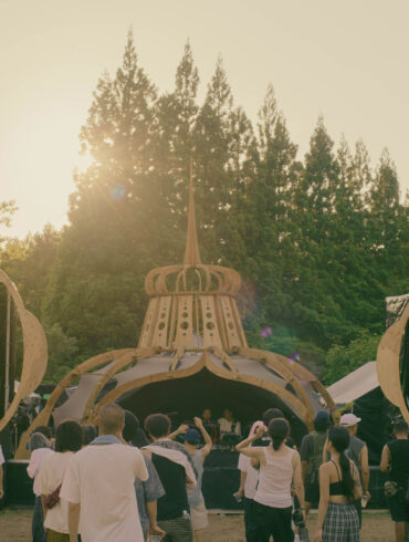 Golden hour at rural 2026 main stage with warm sunset glow and deep listening crowd immersed in techno, set against the forests of Fukushima, Japan