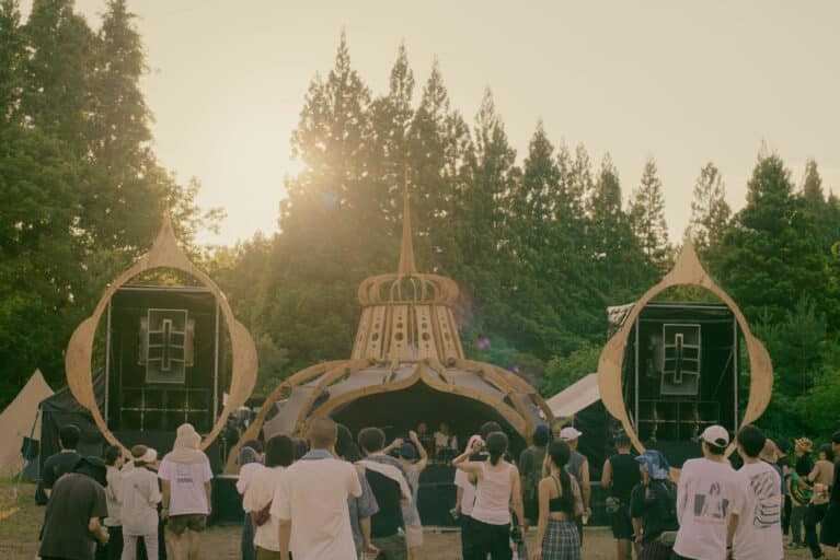 Golden hour at rural 2026 main stage with warm sunset glow and deep listening crowd immersed in techno, set against the forests of Fukushima, Japan