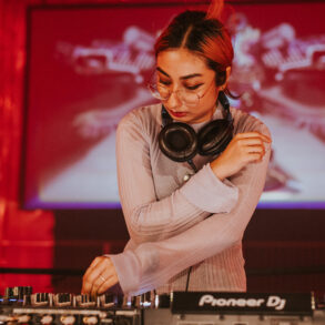 DJ adjusting mixer controls during a live set, wearing headphones around the neck, with red lighting and visuals in the background.