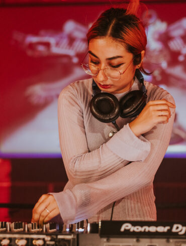 DJ adjusting mixer controls during a live set, wearing headphones around the neck, with red lighting and visuals in the background.