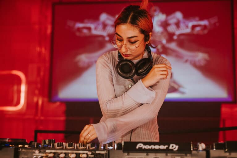 DJ adjusting mixer controls during a live set, wearing headphones around the neck, with red lighting and visuals in the background.