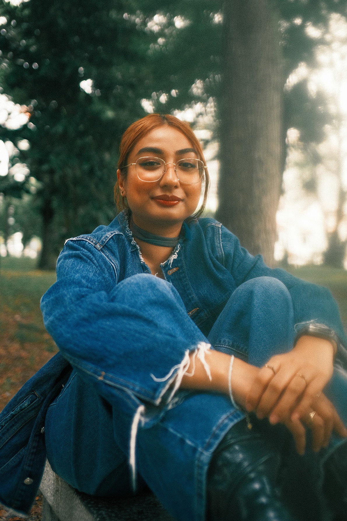 Portrait of a woman seated outdoors in a park, wearing a denim jacket and glasses, looking calmly at the camera in natural light.