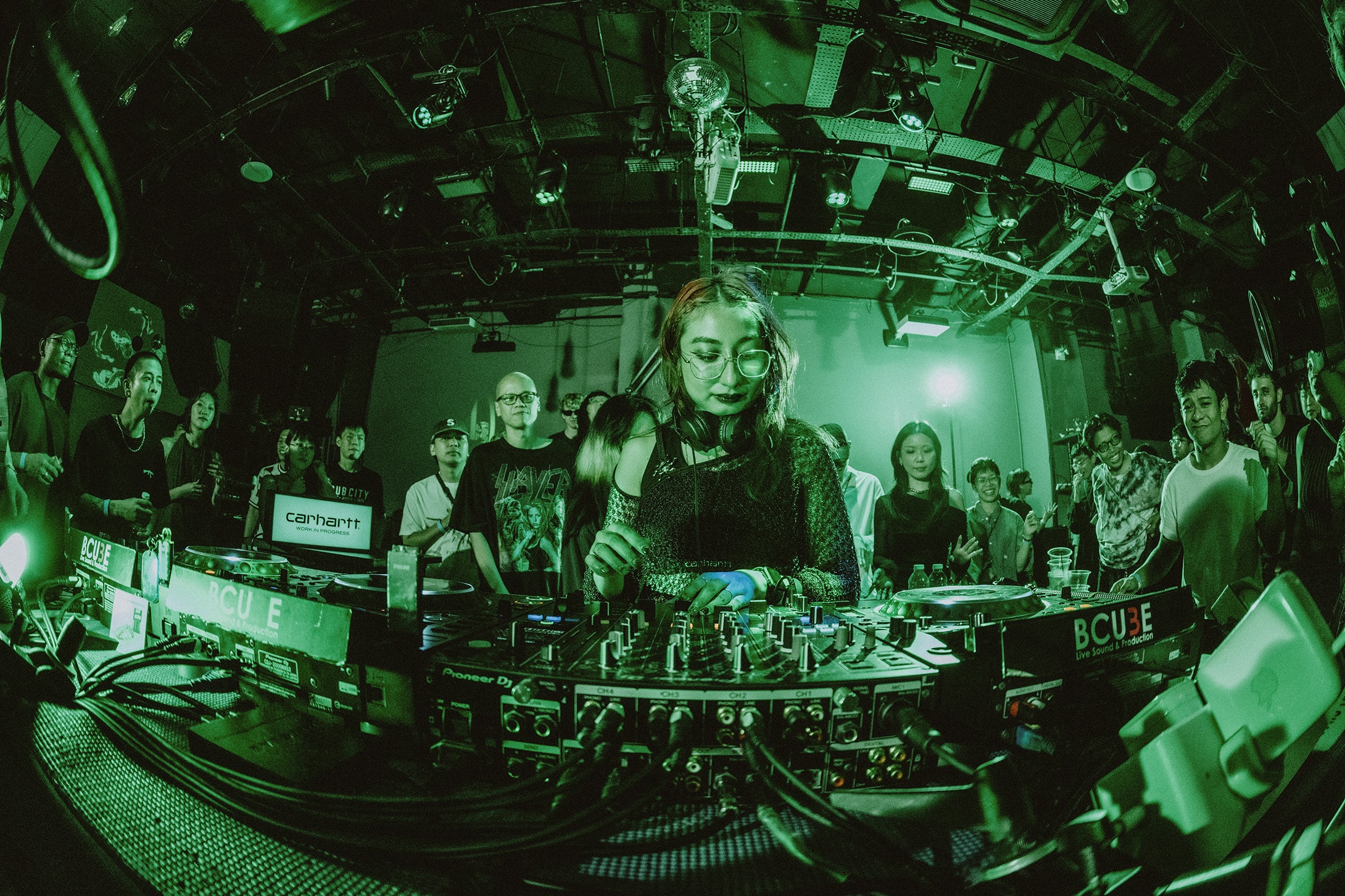 DJ performing at a crowded club venue under green lighting, standing behind a mixer with an audience gathered closely around the booth.
