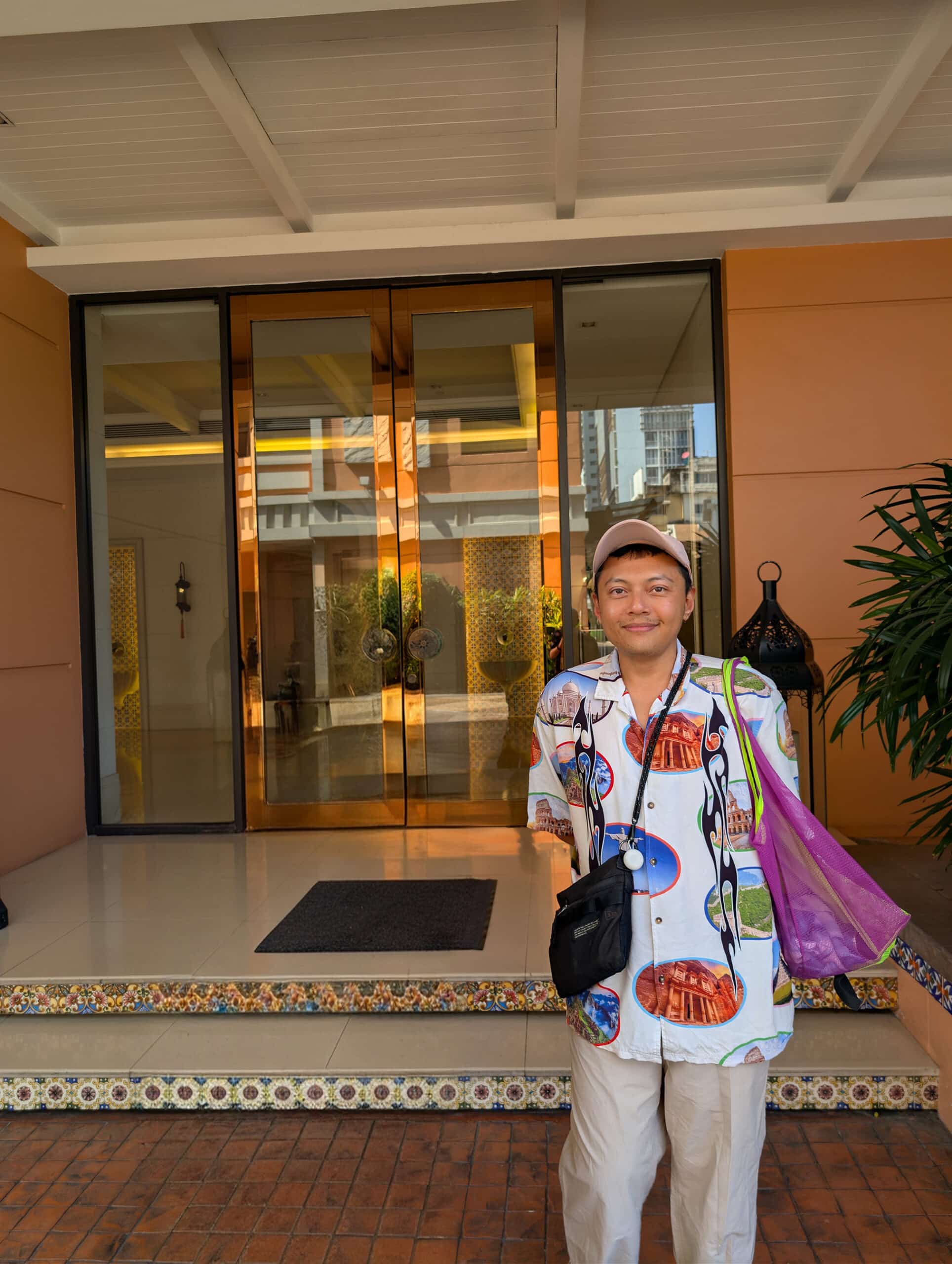 Singaporean Muslim artist Reza Hasni standing outside a building lobby, wearing a patterned shirt and carrying a purple tote bag.