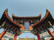 Low-angle view of an ornate traditional Chinese temple gate with curved tiled roofs, red pillars, intricate carvings, and a blue plaque against a clear sky.