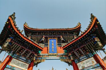 Low-angle view of an ornate traditional Chinese temple gate with curved tiled roofs, red pillars, intricate carvings, and a blue plaque against a clear sky.